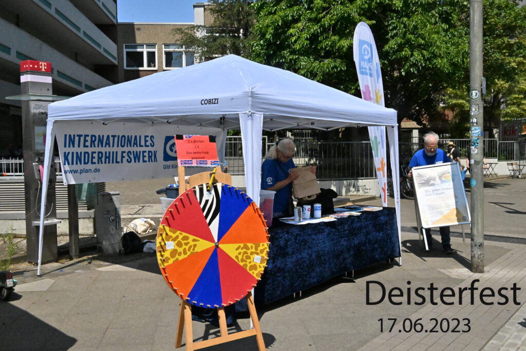 Ein Stand mit einem weißen Vordach, auf dem „Internationales Kinderhilfswerk“ steht, auf dem Deisterfest am 17.06.2023. Zwei Personen bauen Materialien auf, und vor dem Tisch steht ein buntes Spinnrad.