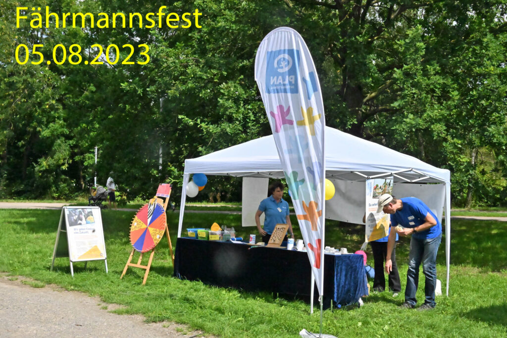 Auf einer Freiluftveranstaltung steht ein weißes Zelt mit einem bunten Banner und einem Spinnrad auf einer Rasenfläche. Zwei Personen betreuen den Stand. Der Text lautet: Fährmannsfest 05.08.2023. Im Hintergrund sind grüne Bäume zu sehen.