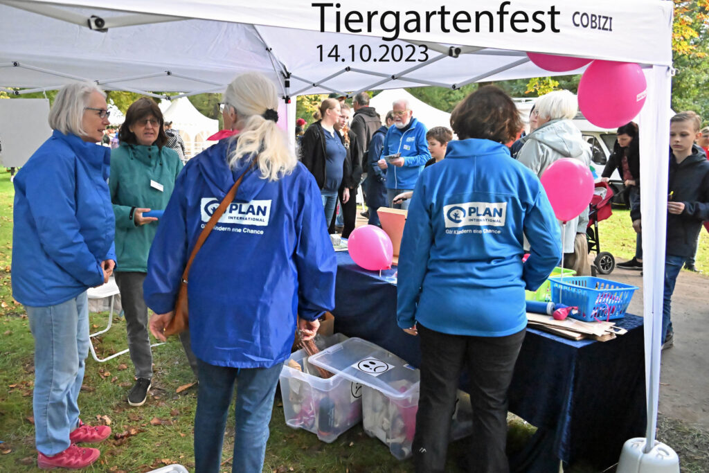 Am 14.10.2023 versammeln sich die Menschen beim Tiergartenfest unter einem weißen Zelt. Freiwillige von Plan International in blauen Jacken stehen den Besuchern zur Seite. Auf dem Tisch liegen rosa Luftballons und Informationsmaterialien.