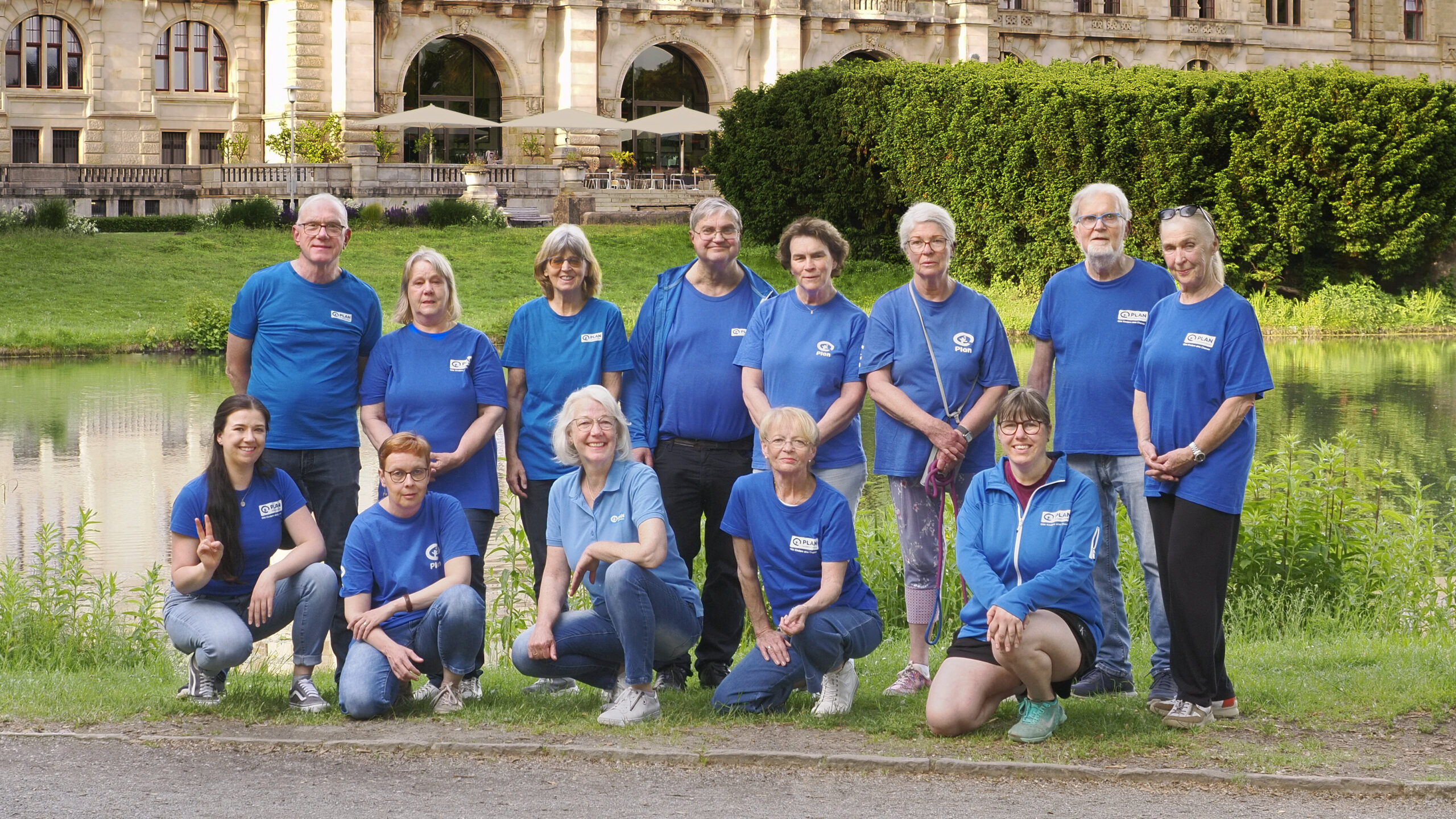 Eine Gruppe von vierzehn Personen in blauen Hemden posiert lächelnd im Freien vor einem historischen Steingebäude mit großen Fenstern, neben einem Teich und inmitten von Grünanlagen.