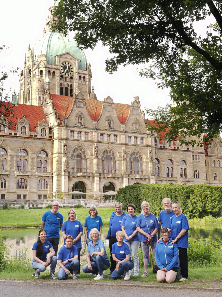 Eine Gruppe von Menschen in blauen Hemden posiert gemeinsam im Freien vor einem großen historischen Gebäude mit einer grünen Kuppel, einem roten Dach und kunstvoller Architektur, im Vordergrund sind Bäume und ein Teich zu sehen.
