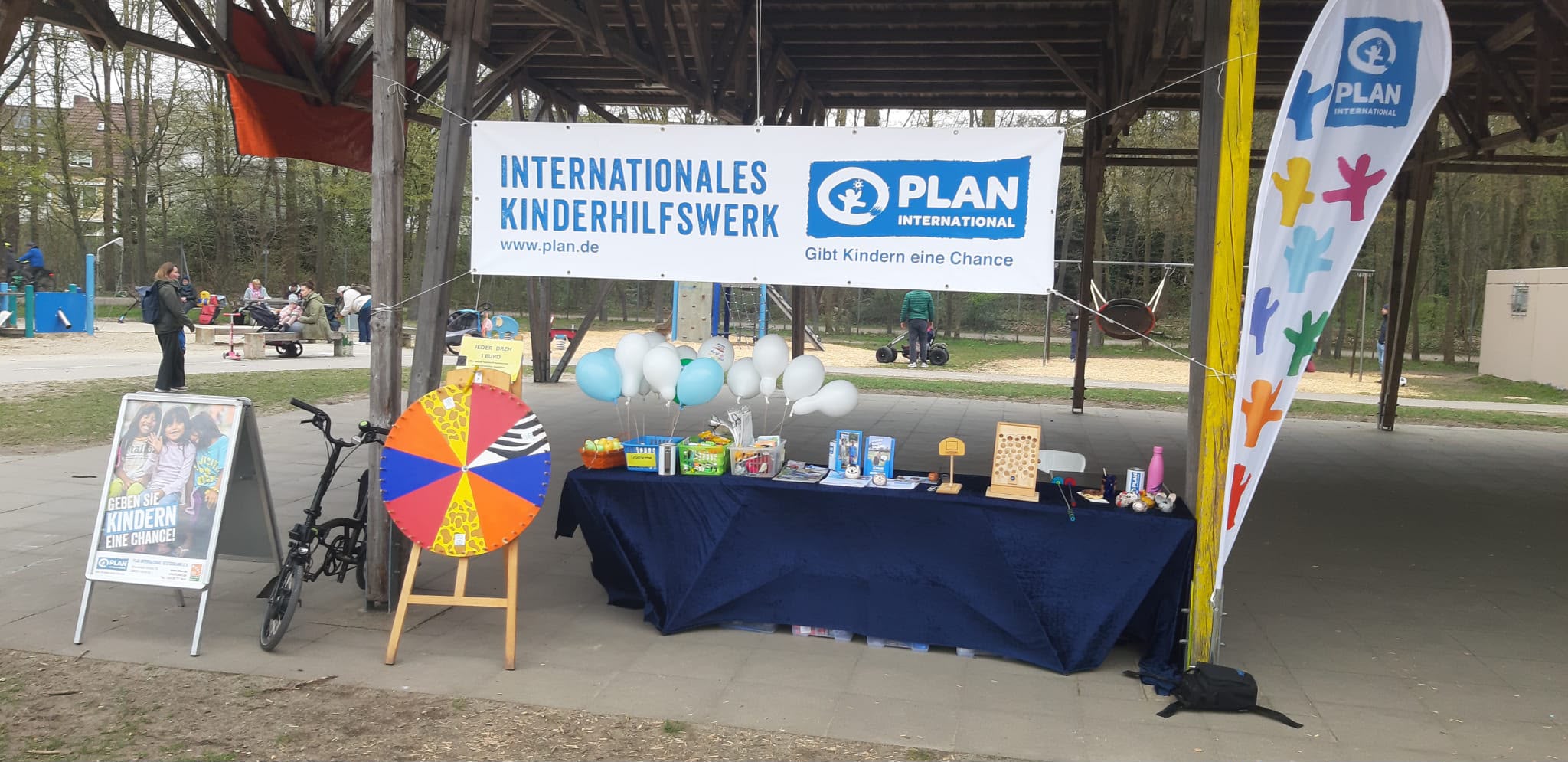 Ein Stand von Plan International mit einem Banner, Informationsflyern, Snacks, Luftballons, einem Glücksrad und einem Ausstellungs-Tisch, der im Freien unter einem Vordach aufgebaut ist. Im Hintergrund sind Menschen zu sehen.