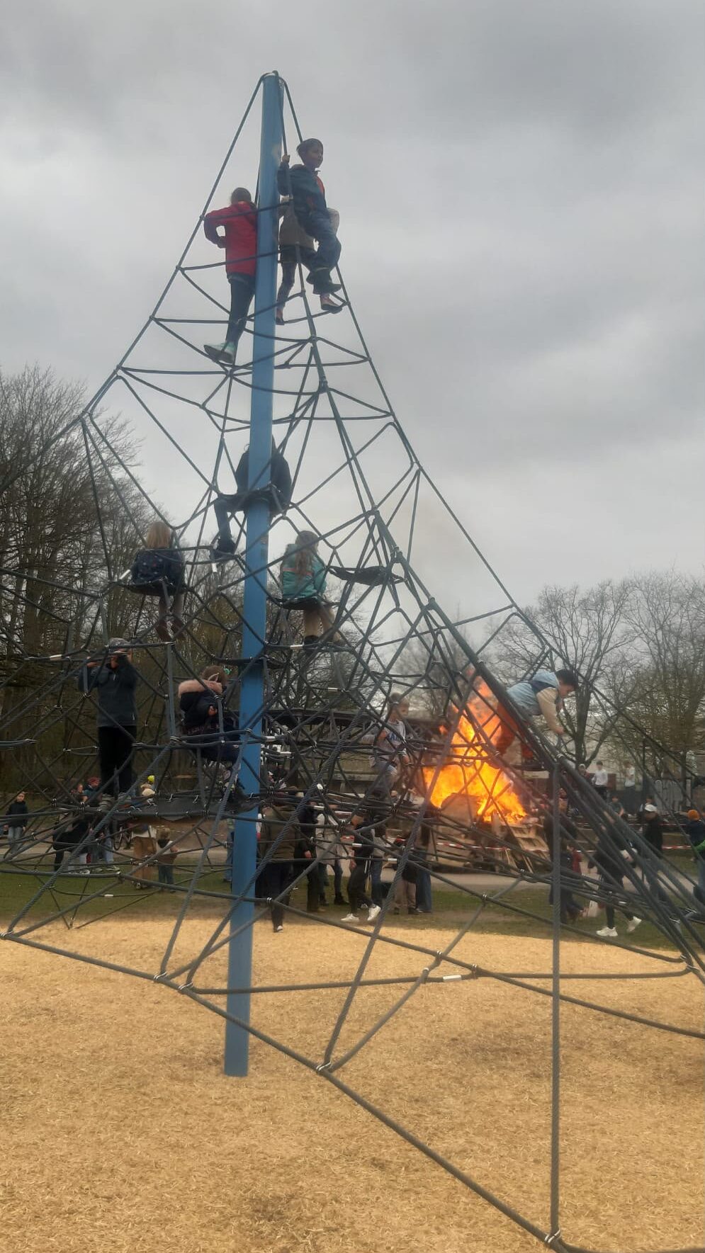 Kinder klettern auf einem Spielplatz auf eine hohe Seilpyramide, während in der Nähe auf dem Boden ein Feuer brennt. Der Himmel ist bewölkt, und im Hintergrund sind Bäume zu sehen.