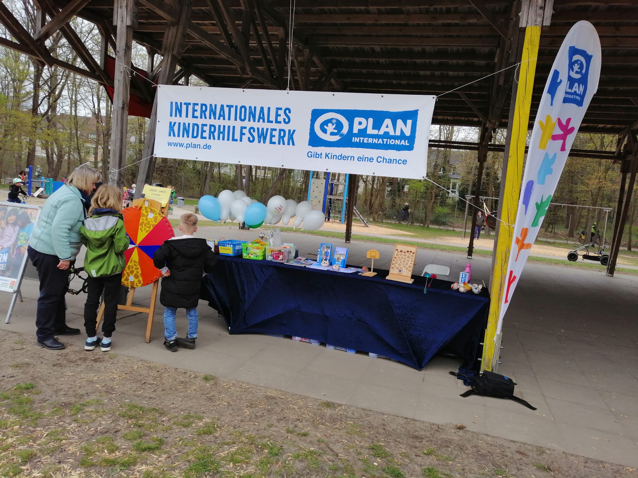 Ein Stand des Plan International Kinderhilfswerks mit Spielen, Spielzeug und Luftballons. Zwei Kinder und ein Erwachsener stehen unter einem Vordach in einem Park neben einem bunten Drehrad.