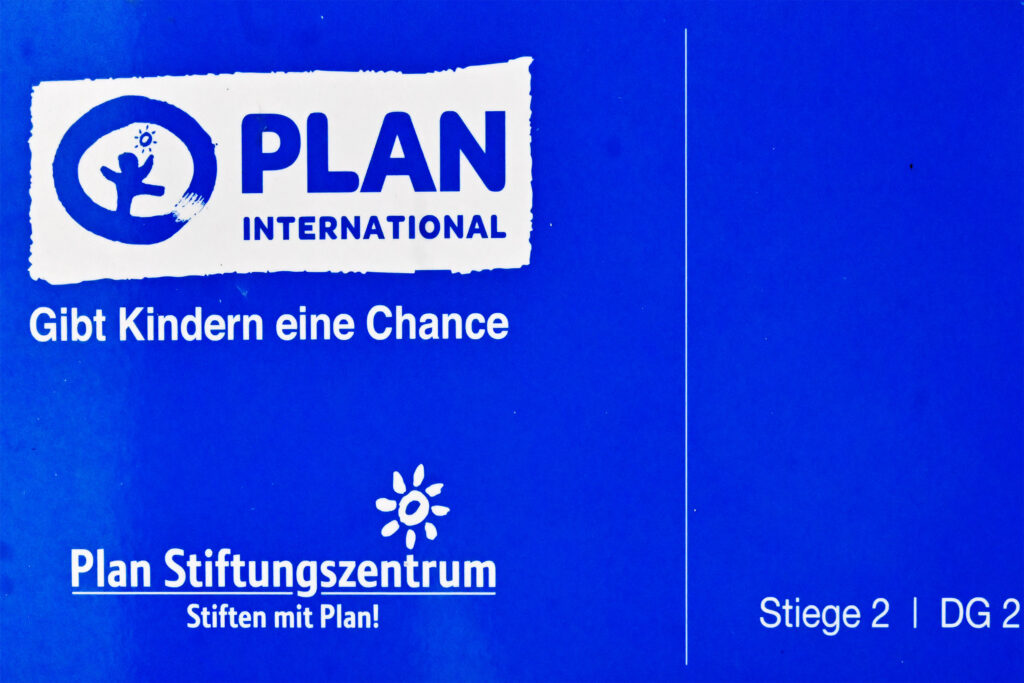 Blaues Schild mit dem PLAN INTERNATIONAL-Logo, den Aufschriften „Gibt Kindern eine Chance“, „Plan Stiftungszentrum – Stiften mit Plan!“ sowie folgenden Standortangaben: Stiege 2 | DG 2.