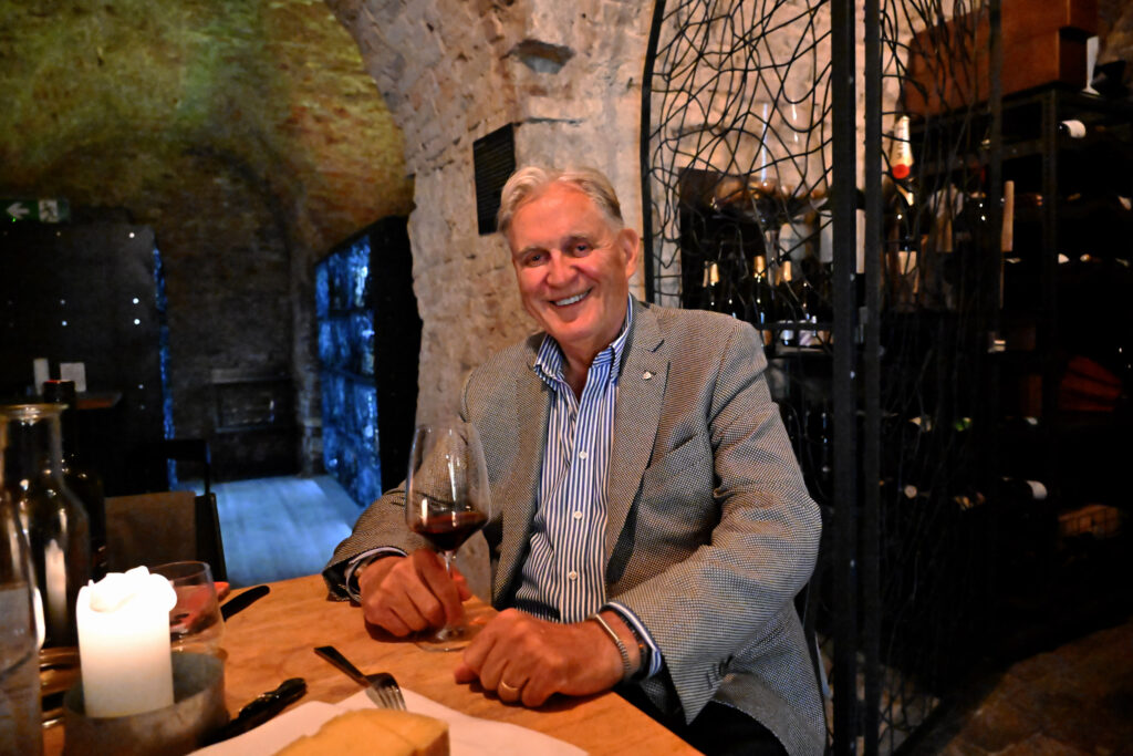 Ein älterer Mann in einem grauen Blazer sitzt an einem Tisch in einem rustikalen Weinkeller, lächelt und hält ein Glas Rotwein in der Hand. Auf dem Tisch und in den Regalen hinter ihm sind eine brennende Kerze, Käse und Weinflaschen zu sehen.